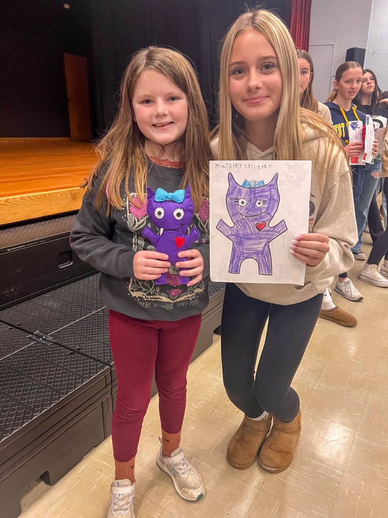 A Goshen High School student and Goshen Intermediate Student pose together. The high school student holds the drawing, and the intermediate student holds the sewn monster they collaborated on. 