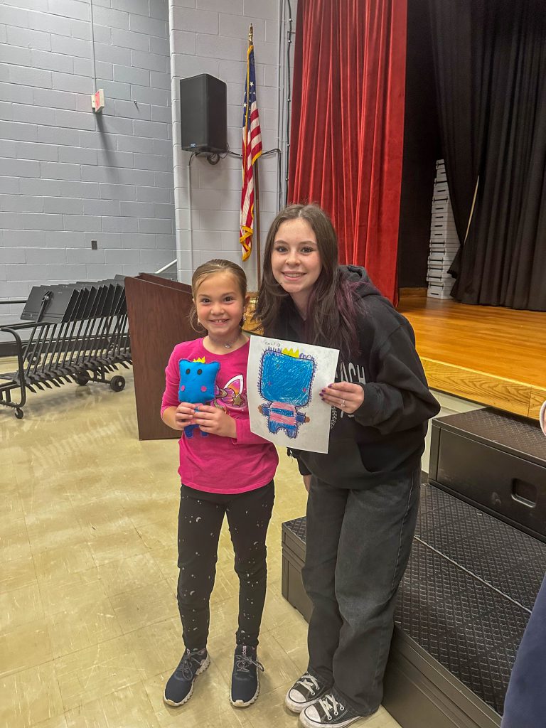 A Goshen High School student and Goshen Intermediate Student pose together. The high school student holds the drawing, and the intermediate student holds the sewn monster they collaborated on. 