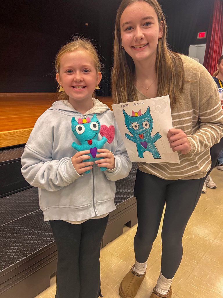 A Goshen High School student and Goshen Intermediate Student pose together. The high school student holds the drawing, and the intermediate student holds the sewn monster they collaborated on. 