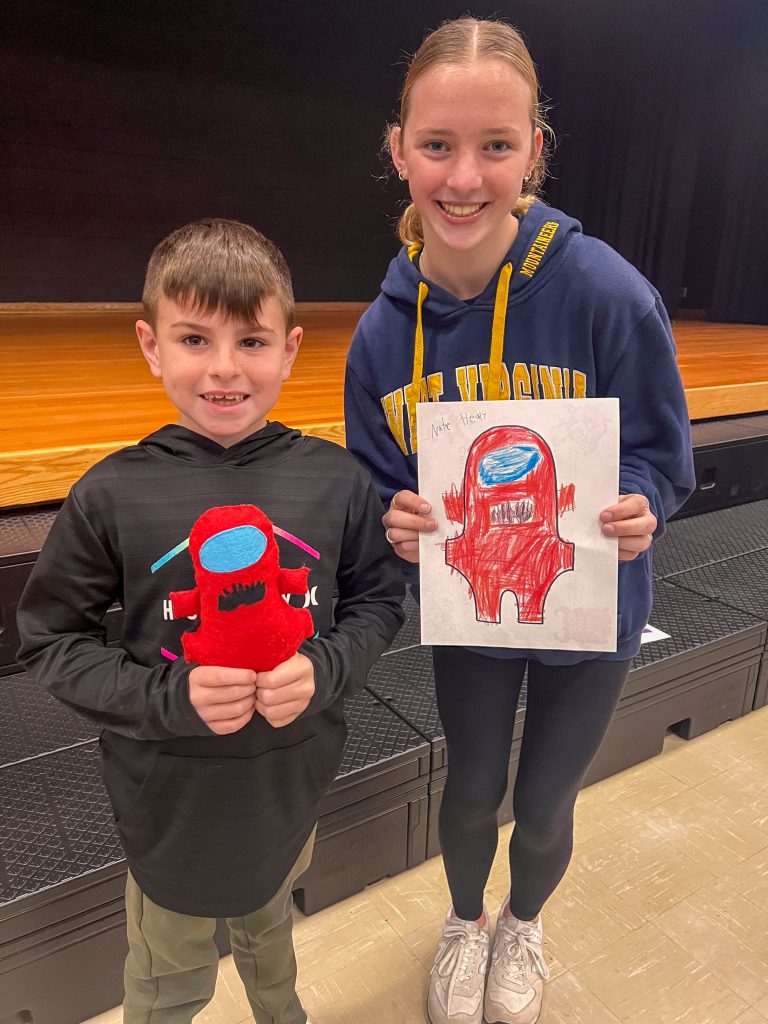 A Goshen High School student and Goshen Intermediate Student pose together. The high school student holds the drawing, and the intermediate student holds the sewn monster they collaborated on. 