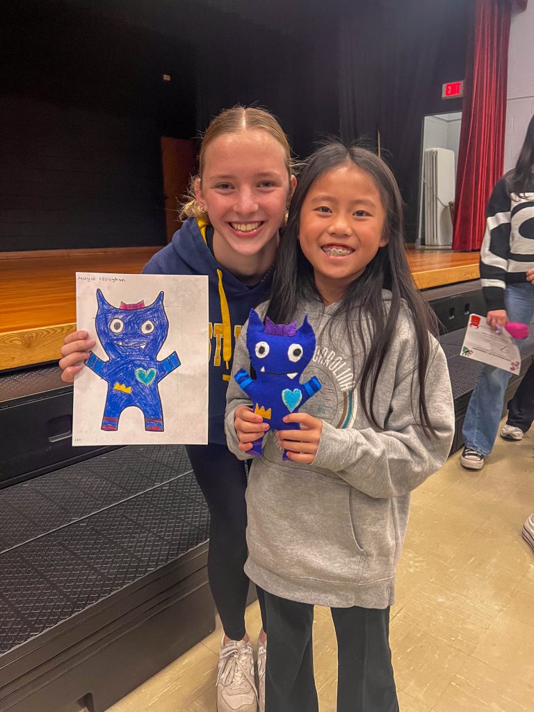 A Goshen High School student and Goshen Intermediate Student pose together. The high school student holds the drawing, and the intermediate student holds the sewn monster they collaborated on. 