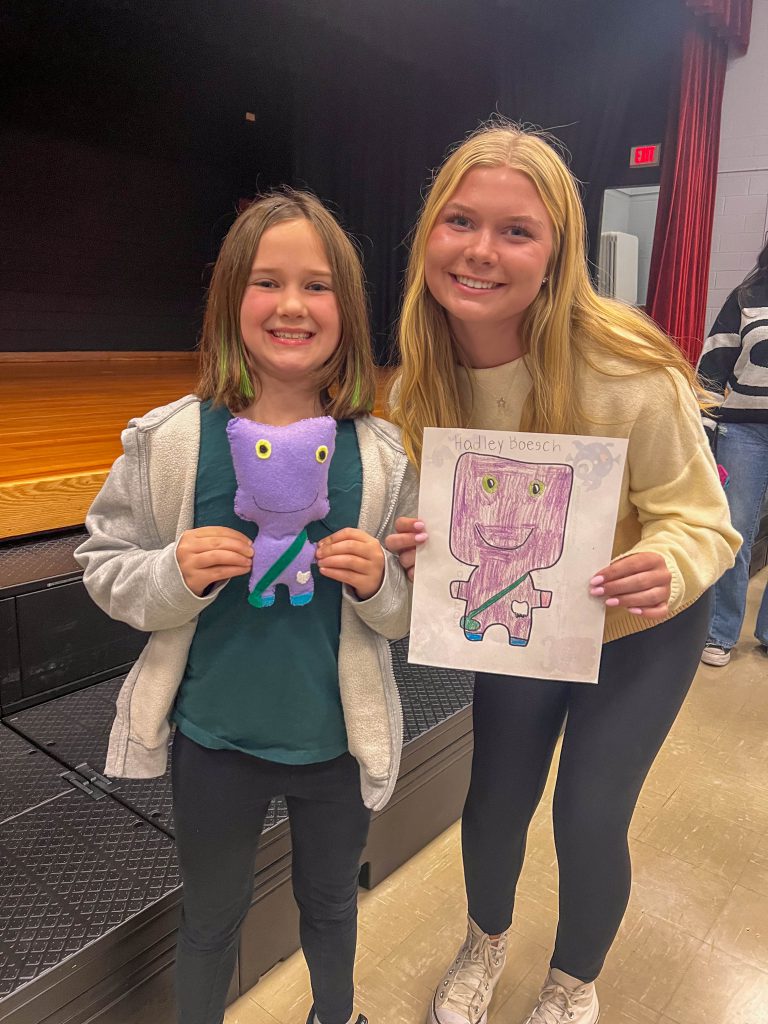 A Goshen High School student and Goshen Intermediate Student pose together. The high school student holds the drawing, and the intermediate student holds the sewn monster they collaborated on. 