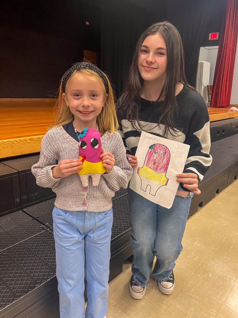 A Goshen High School student and Goshen Intermediate Student pose together. The high school student holds the drawing, and the intermediate student holds the sewn monster they collaborated on. 