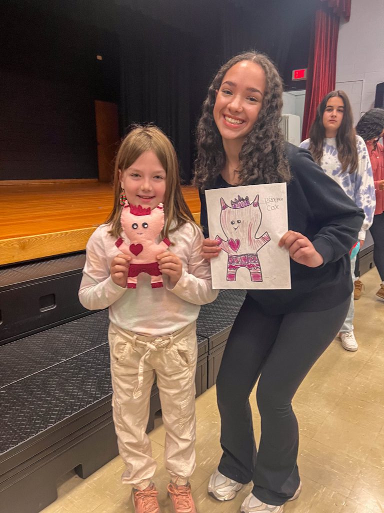 A Goshen High School student and Goshen Intermediate Student pose together. The high school student holds the drawing, and the intermediate student holds the sewn monster they collaborated on. 