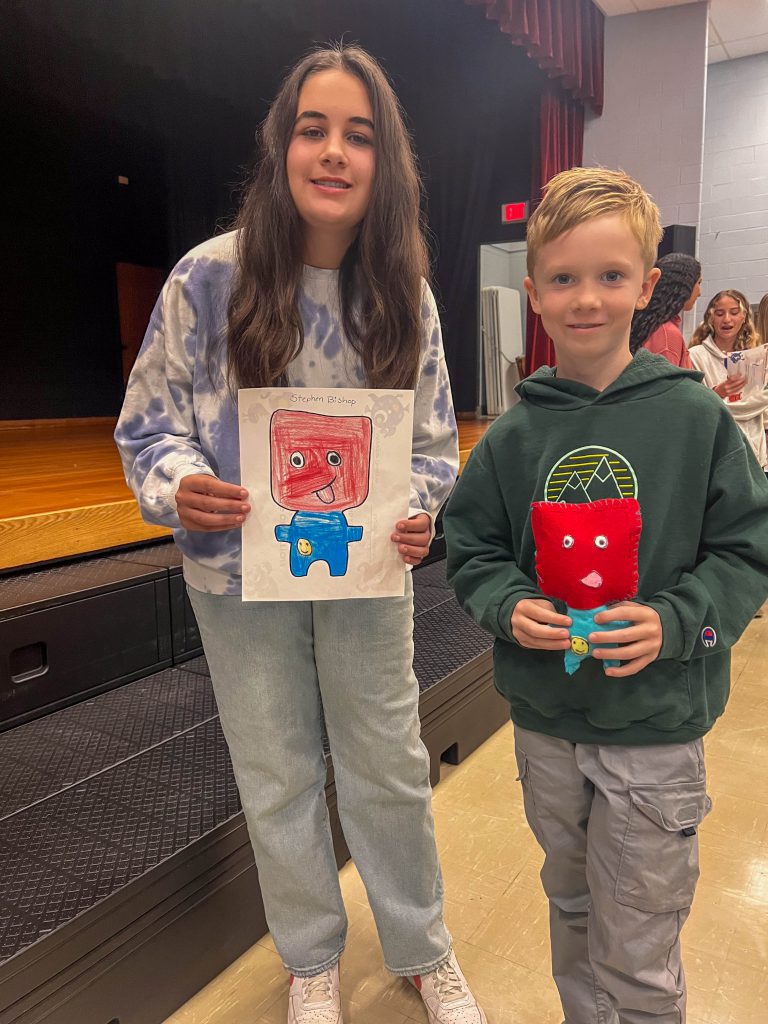 A Goshen High School student and Goshen Intermediate Student pose together. The high school student holds the drawing, and the intermediate student holds the sewn monster they collaborated on. 