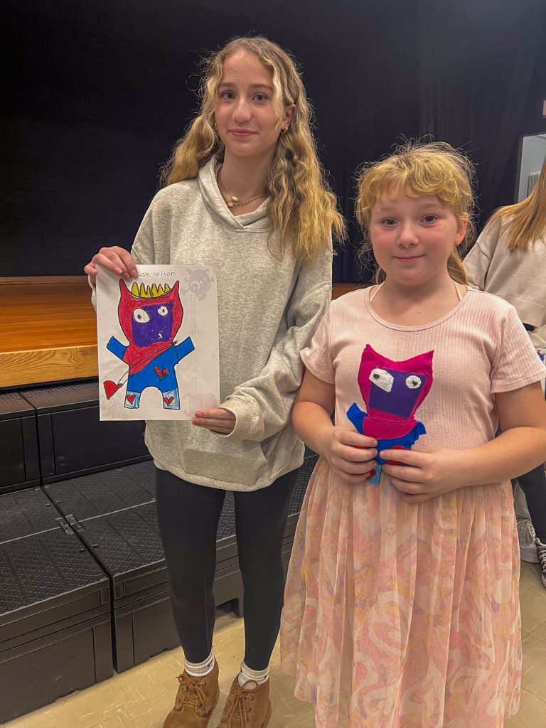 A Goshen High School student and Goshen Intermediate Student pose together. The high school student holds the drawing, and the intermediate student holds the sewn monster they collaborated on. 