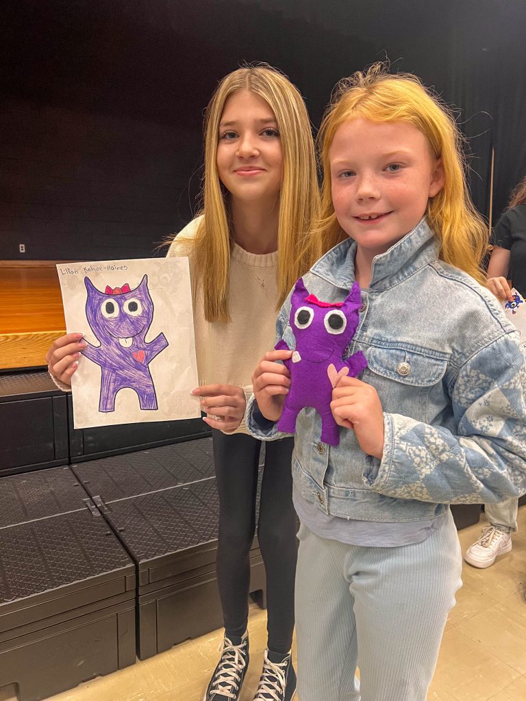 A Goshen High School student and Goshen Intermediate Student pose together. The high school student holds the drawing, and the intermediate student holds the sewn monster they collaborated on. 