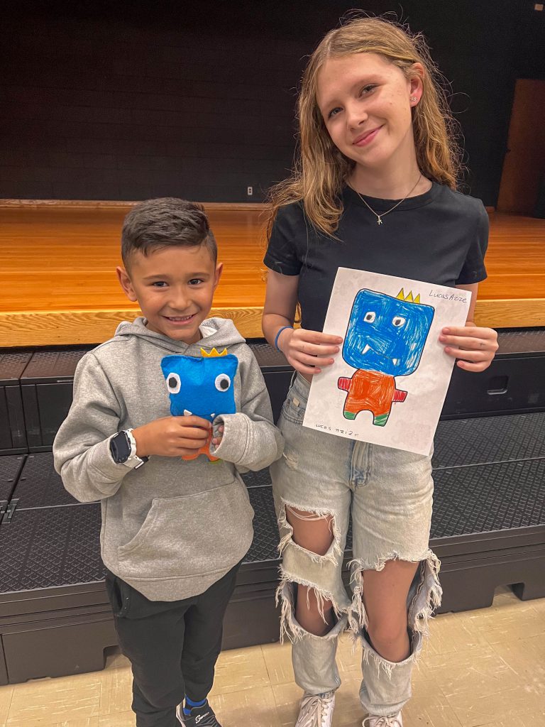 A Goshen High School student and Goshen Intermediate Student pose together. The high school student holds the drawing, and the intermediate student holds the sewn monster they collaborated on. 