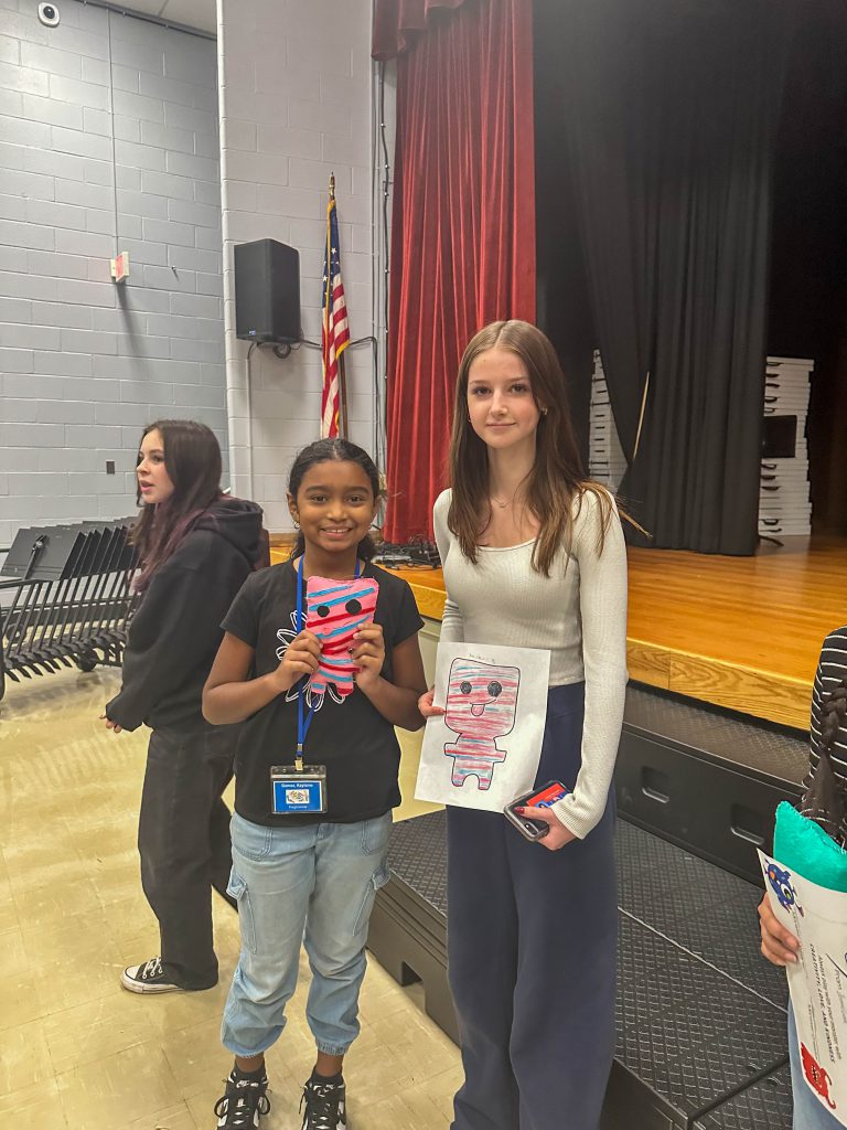 A Goshen High School student and Goshen Intermediate Student pose together. The high school student holds the drawing, and the intermediate student holds the sewn monster they collaborated on. 