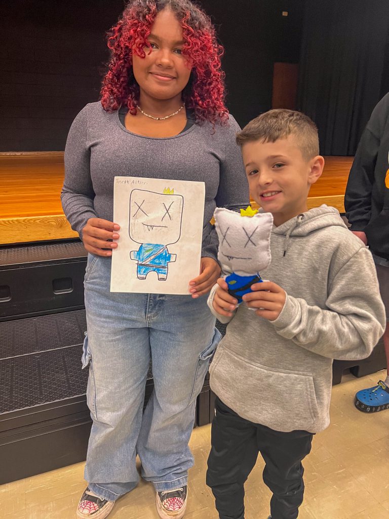 A Goshen High School student and Goshen Intermediate Student pose together. The high school student holds the drawing, and the intermediate student holds the sewn monster they collaborated on. 