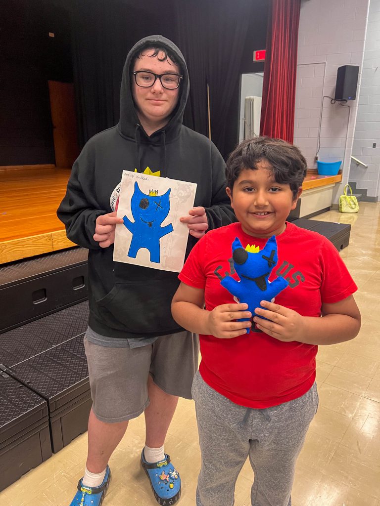 A Goshen High School student and Goshen Intermediate Student pose together. The high school student holds the drawing, and the intermediate student holds the sewn monster they collaborated on. 