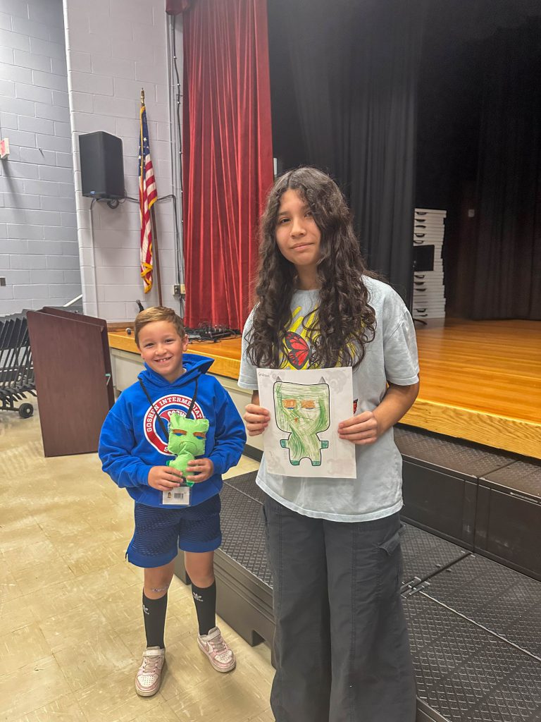 A Goshen High School student and Goshen Intermediate Student pose together. The high school student holds the drawing, and the intermediate student holds the sewn monster they collaborated on. 