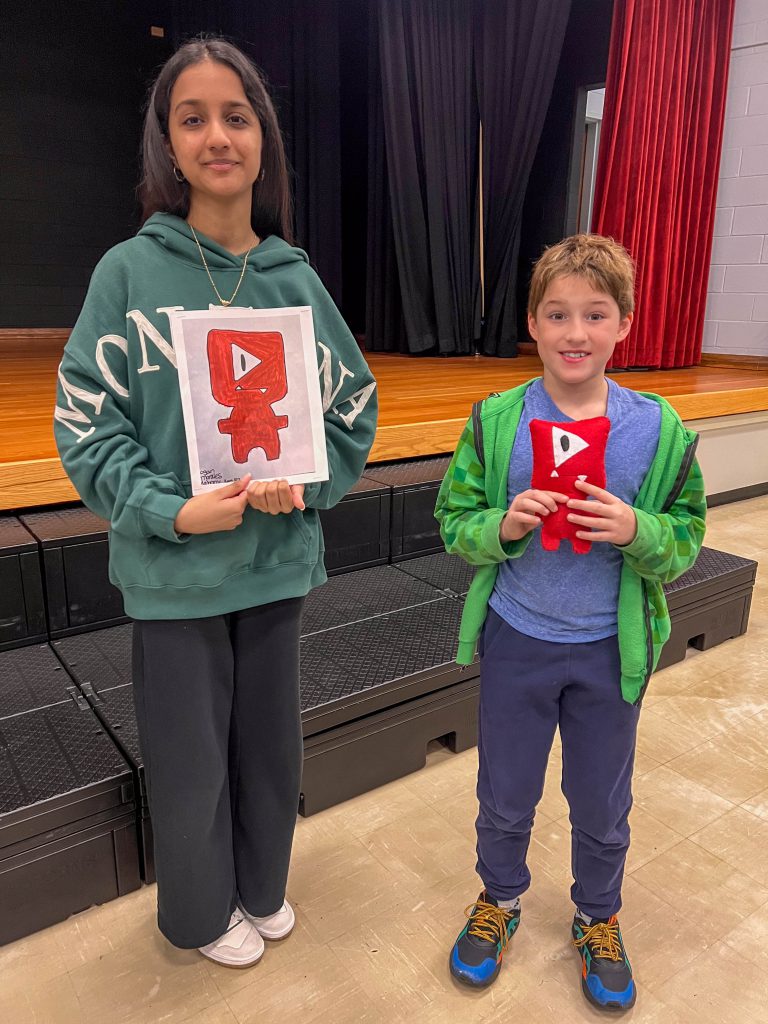 A Goshen High School student and Goshen Intermediate Student pose together. The high school student holds the drawing, and the intermediate student holds the sewn monster they collaborated on. 