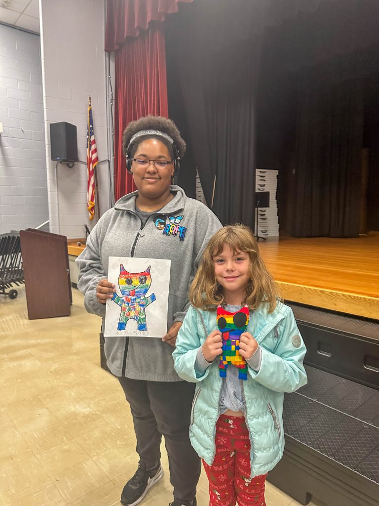 A Goshen High School student and Goshen Intermediate Student pose together. The high school student holds the drawing, and the intermediate student holds the sewn monster they collaborated on. 