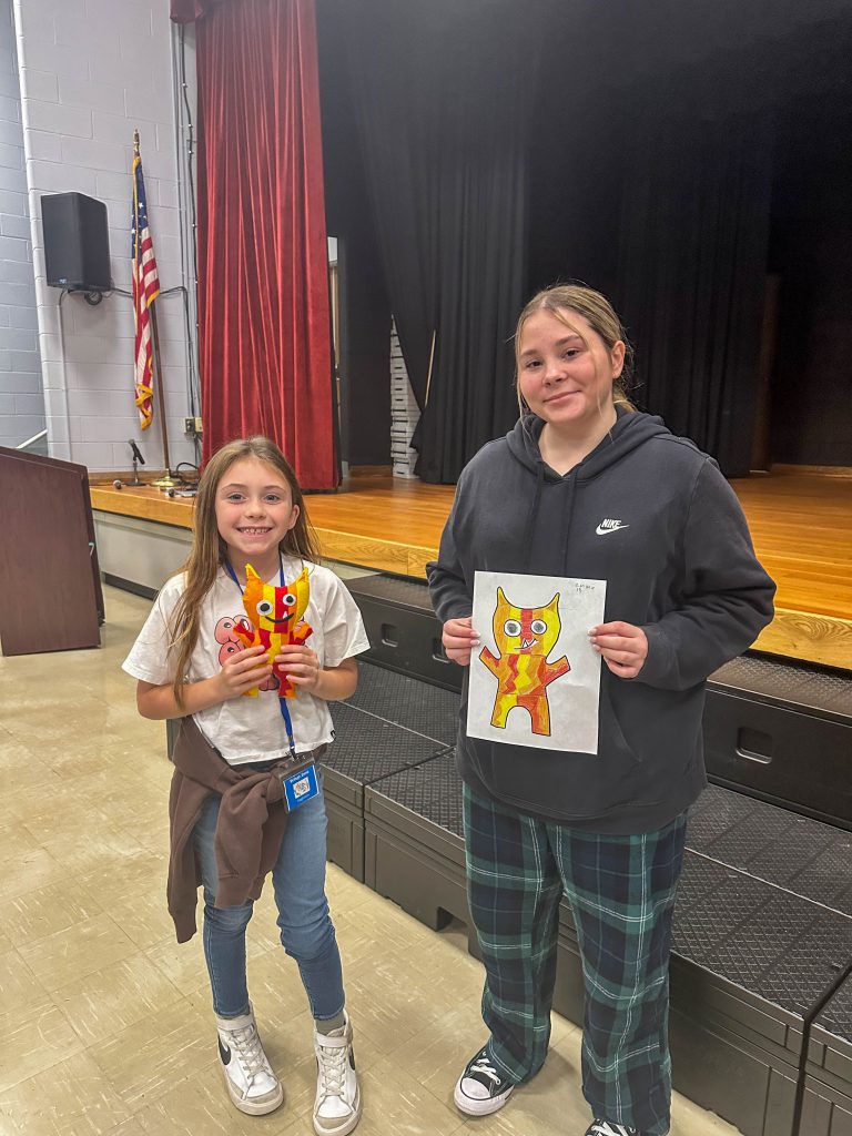 A Goshen High School student and Goshen Intermediate Student pose together. The high school student holds the drawing, and the intermediate student holds the sewn monster they collaborated on. 