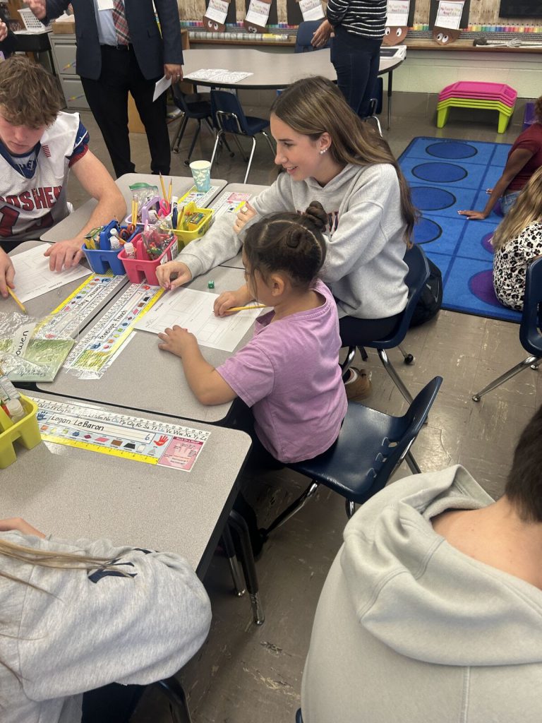 A Goshen High School student sits with a Scotchtown Avenue students as they discuss some school work and fill out a worksheet. The table is filled with students from each school working together. 