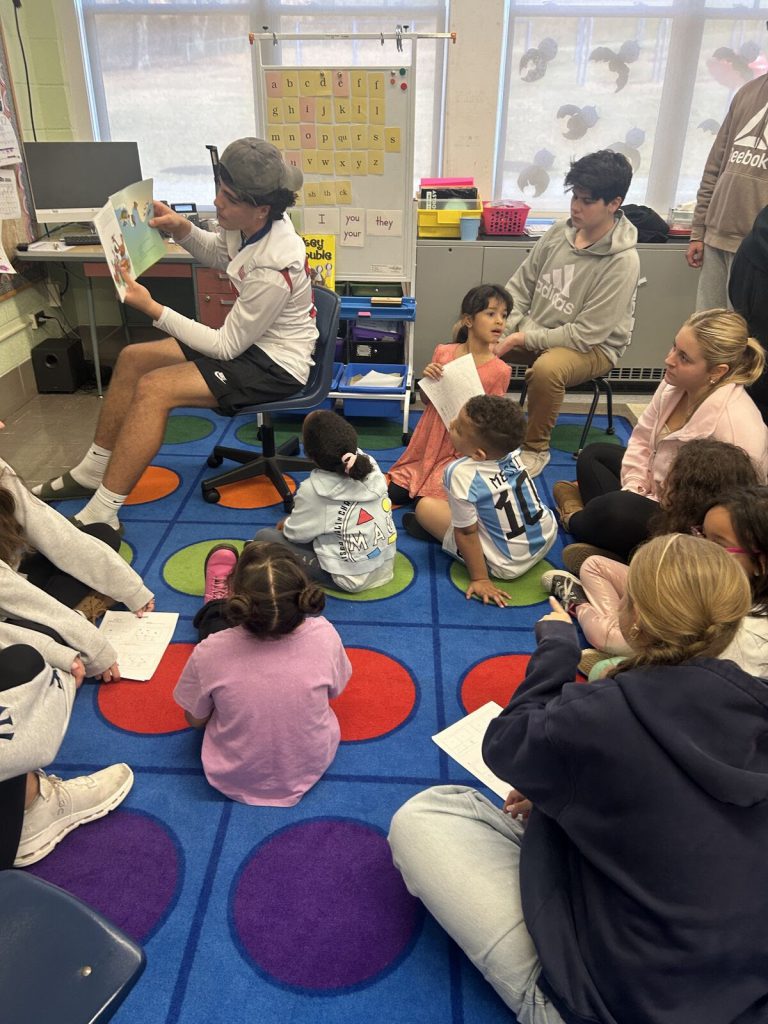 A Goshen High School student reads a book to Scotchtown Avenue students sitting on the rug. The student reading is holding the picture book over their shoulder so that the students can see from the rug. 