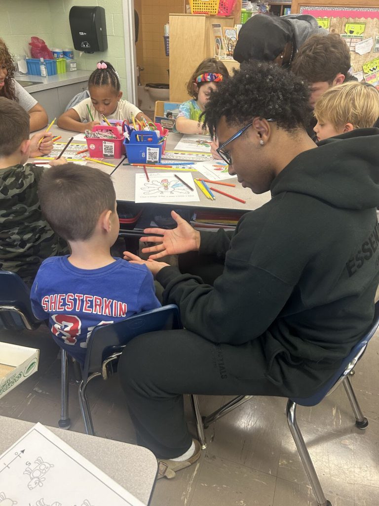 A Goshen High School student is working with a Scotchtown Avenue student as they color a Thanksgiving turkey on a paper. Students around the table are holding colored pencils and filling in the feathers of their turkey. 