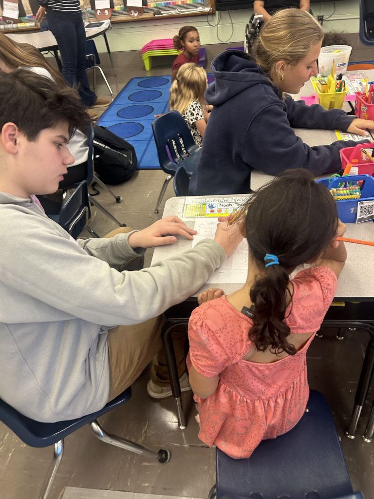 A Goshen High School student sits with a Scotchtown Avenue students as they discuss some school work and fill out a worksheet. Students are sitting on a carpet in the background while they are learning as well.