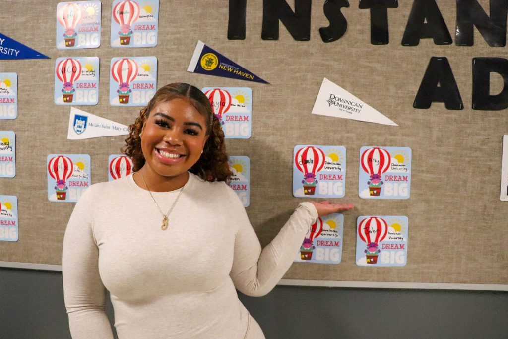 A student stands near her name under an "Dominican University" banner on the wall at the instant admissions board in the hallway at Goshen High School. 