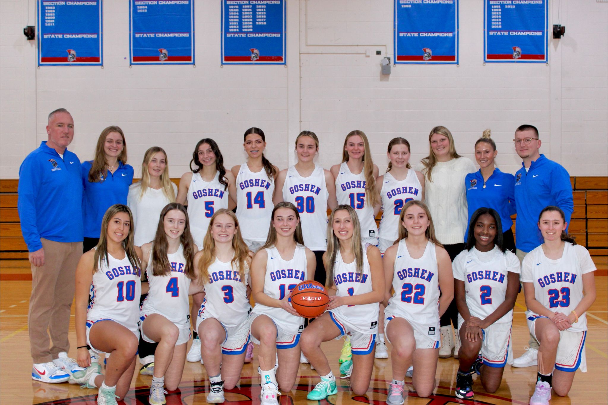 The Goshen Girls Varsity basketball team poses for their team photo. There is a row in front kneeling down and a row behind standing. The coaches are on the sides of the team.