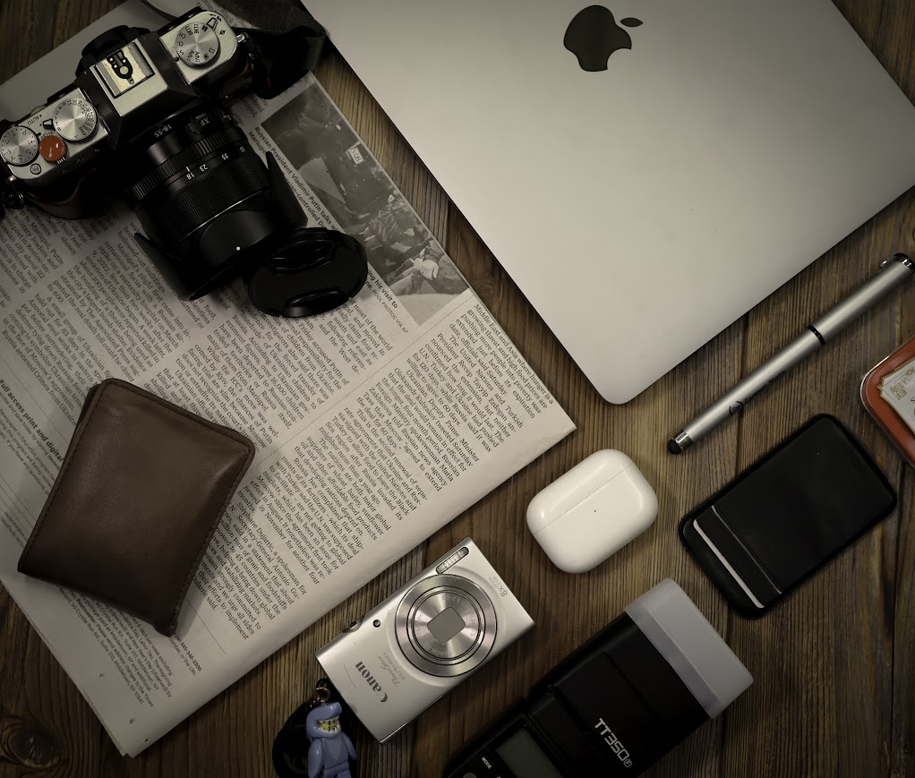 A photo of camera gear, a newspaper, laptop, and other things found on a desk. The photo is taken from above the table these things are on.  