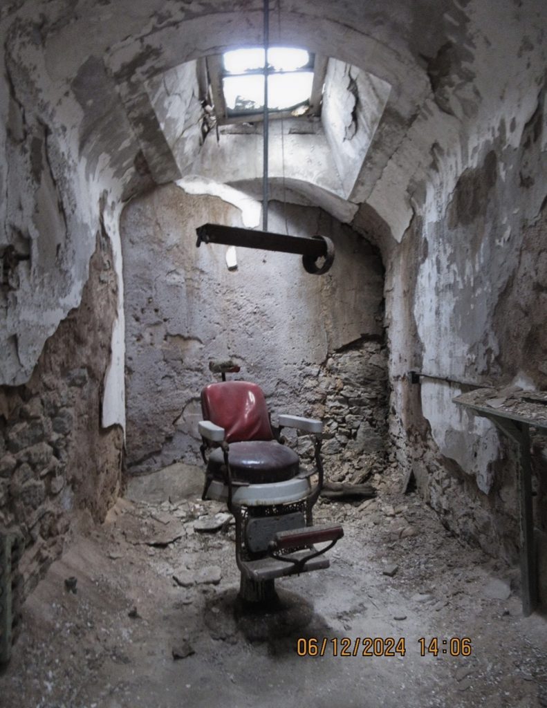 A chair in the middle of a seemingly abandoned room. The room has rocks all over and the concrete walls are exposed beneath a skylight. 