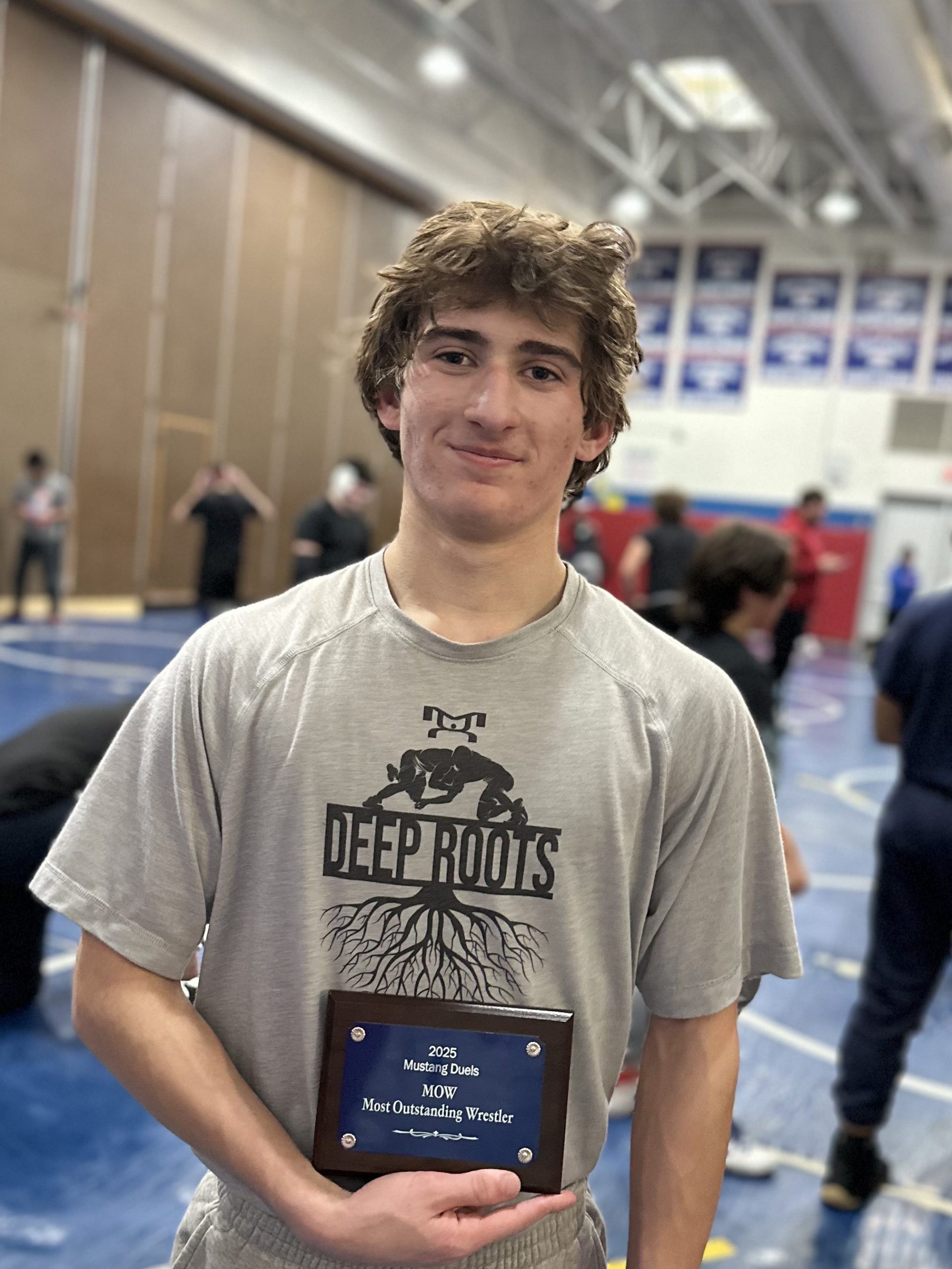 Greg Schaefer stands and holds a plaque that says "Most Outstanding Wrestler" and "2025 Mustang Duals." He is in the gym with others in the background.  