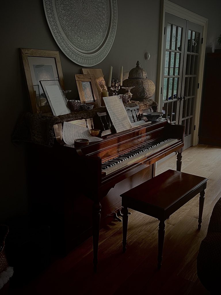 A piano set with music and photos on it. A lot of shadow is around the piano in a room with glass doors on the wall in the background. 