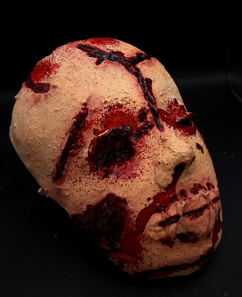 A sculpture of a face with blood and cuts all over it. There are no eyes and the mouth is closed. It is in front of a black background. 