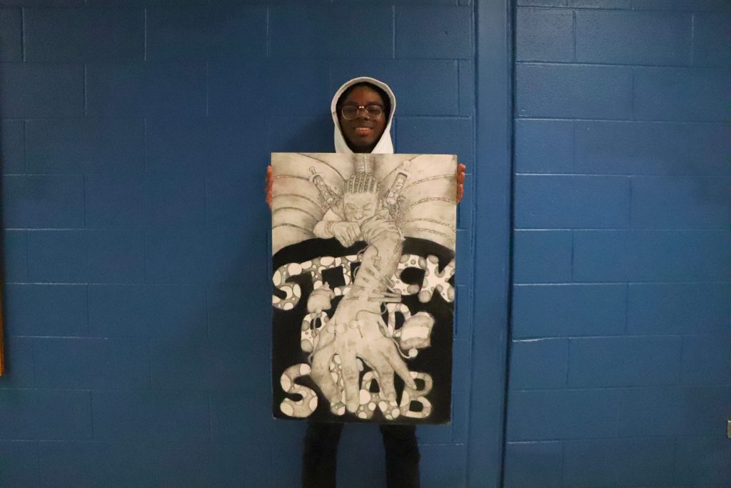 Tahir Bernard holds his work "Stick and Stab" which is a person trying to climb onto a platform with the words stick and stab written on it. There are syringes injected into the person in a black and white photo/ 