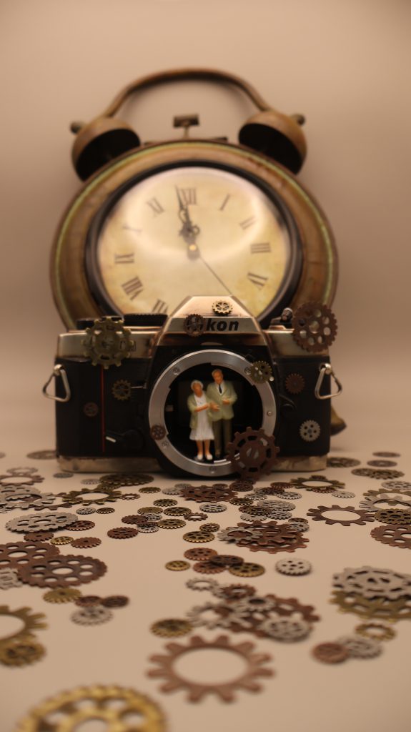 An older couple holds hands inside a camera body with springs from a clock on the floor. Behind them is a large clock that goes on a wall. 