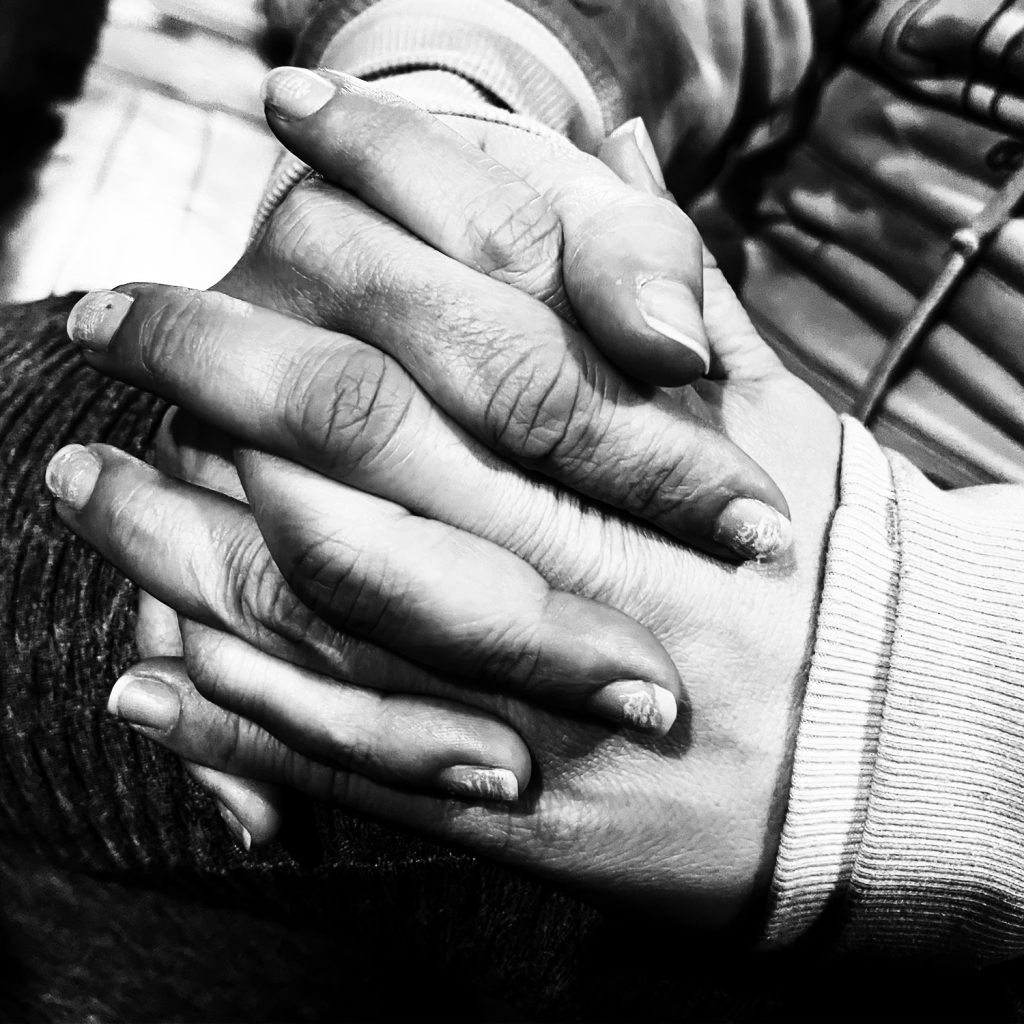 Hands holding each other close up. The finger nails are long and the person is wearing a sweater. The photo is black and white.