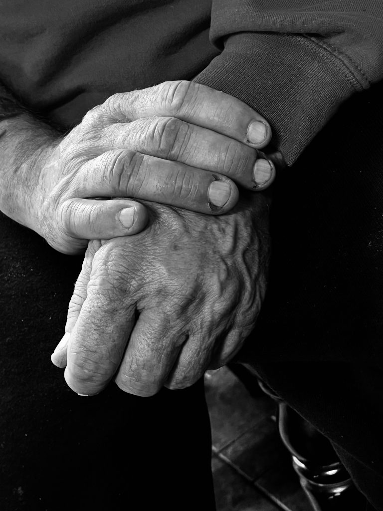 A person holds their left hand in their right. The picture is gray and the hands are wrinkled. 