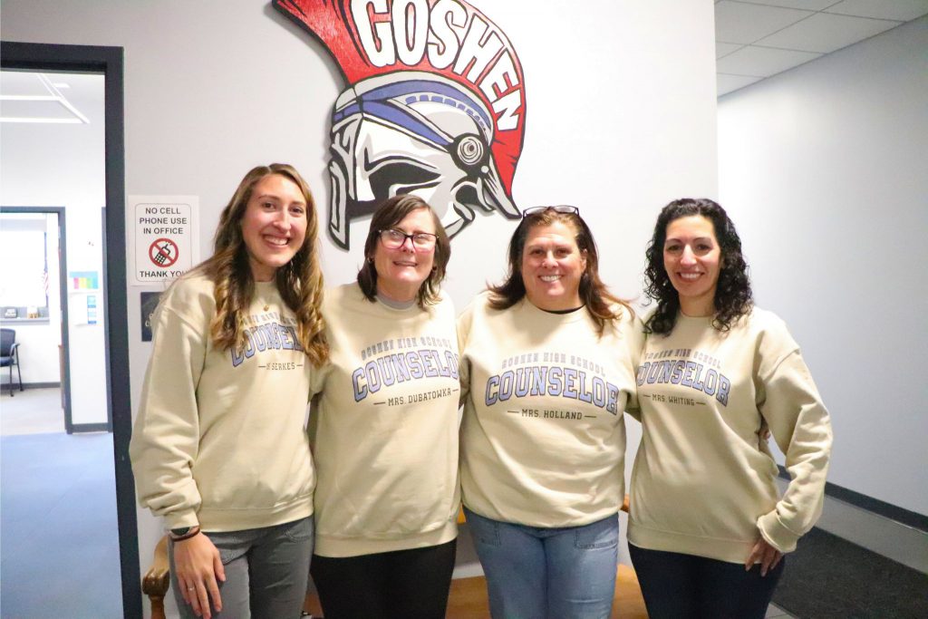 The four Goshen High School Guidance Counselors are standing in a row with customized sweaters. They are in front of the Goshen Gladiator helmet and smiling for the camera. 