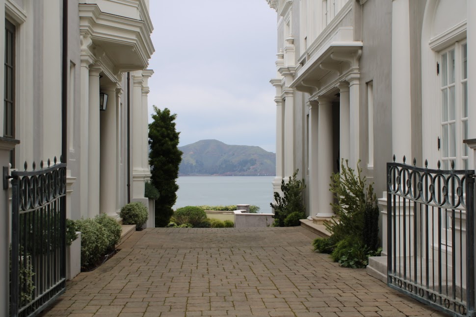 White buildings with columns and a stone floor overlook water with a mountain in the background. The photo is taken through an opening in the buildings. 