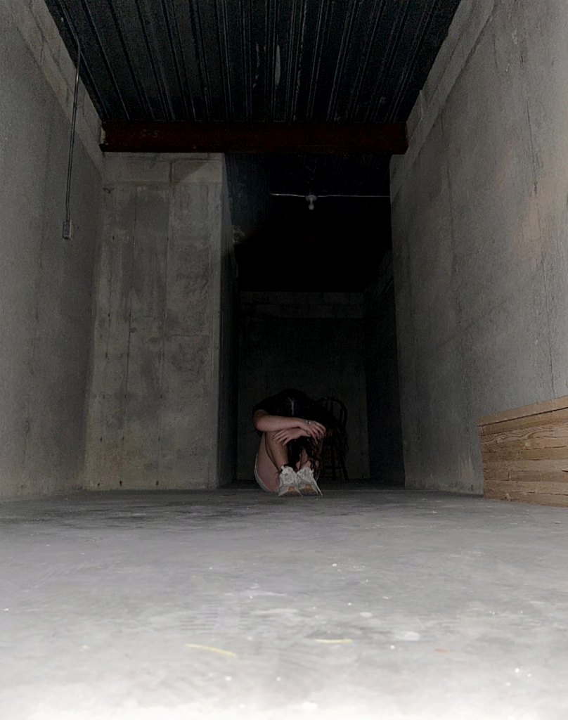 A person is sitting on a cement floor with their arms around their legs in a dark area. The entire structure is cement and it is dark behind the person and above on the ceiling. 