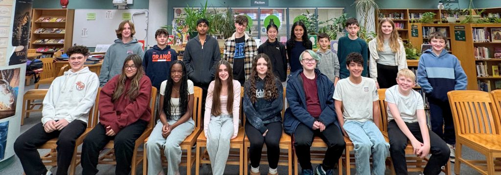 Two rows of students are smiling at the camera. The front row is sitting in chairs and the back row is standing. The students are in the library with books and plants all around them. 