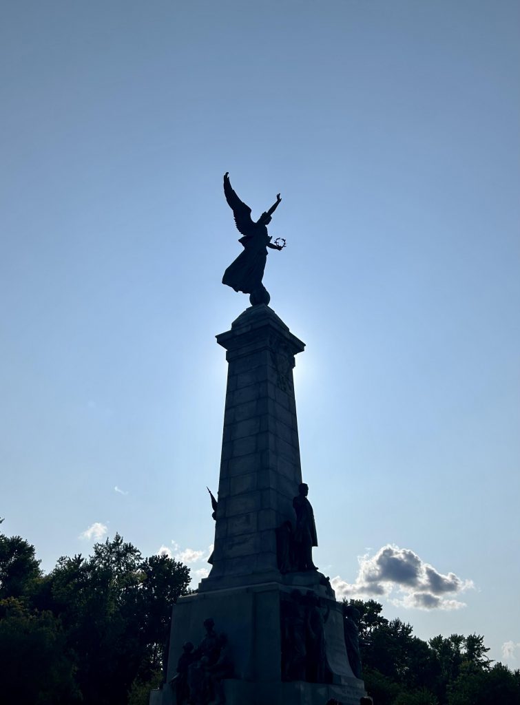 A statue of a person with wings is at the top of a monument with other sculptured people below. The sun is behind the structure and the photo is taken from within the shadow. 