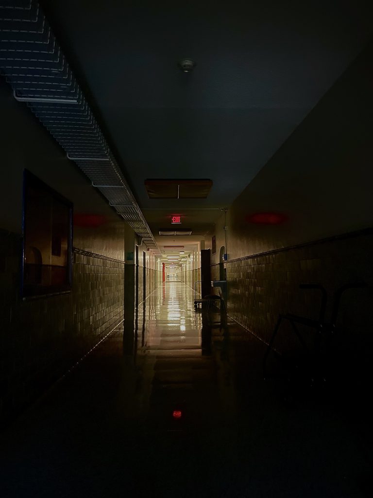 A dark hallway is near the camera while the lights are on in the distance down the hallway. It is in a building with tile walls. 