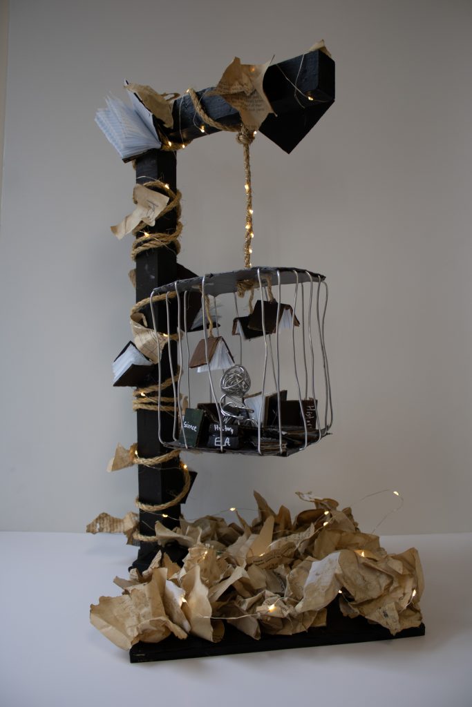 A sculpture of a cage hanging down by a string with lights all over from a structure with books hanging from the ceiling of the cage. There are papers on the ground below the cage. There are books on the structure arms that hold the cage up. 