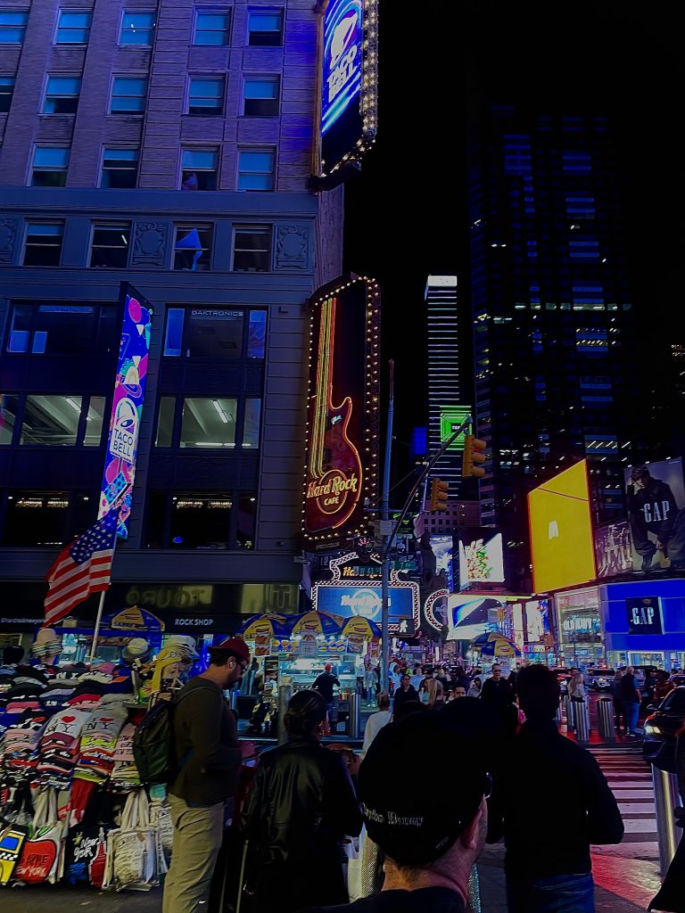 A photo of the Hard Rock Cafe with advertisements and large glowing signs around it. There are people walking in the street and people selling things on the sidewalks. 