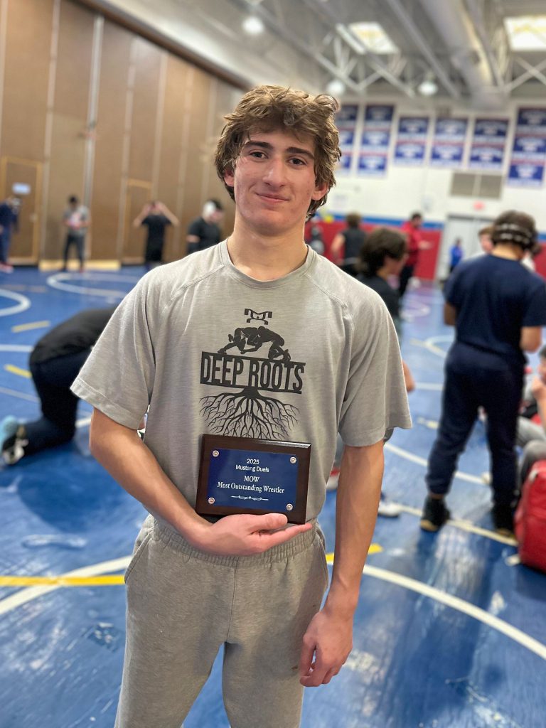 Greg holds a plaque and smiles for the camera at practice. The plaque says 2025 Mustang Duels MOW - Most Outstanding Wrestler."