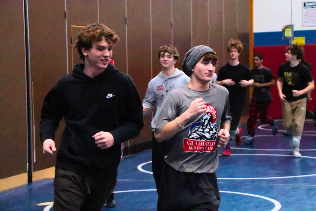 Evan Weeden and Greg Schaefer lead the wrestling team as they run around the gym during warm ups. Evan is on the left and is wearing a black sweater. Greg is on the right and wearing a Goshen Gladiators wrestling shirt. 