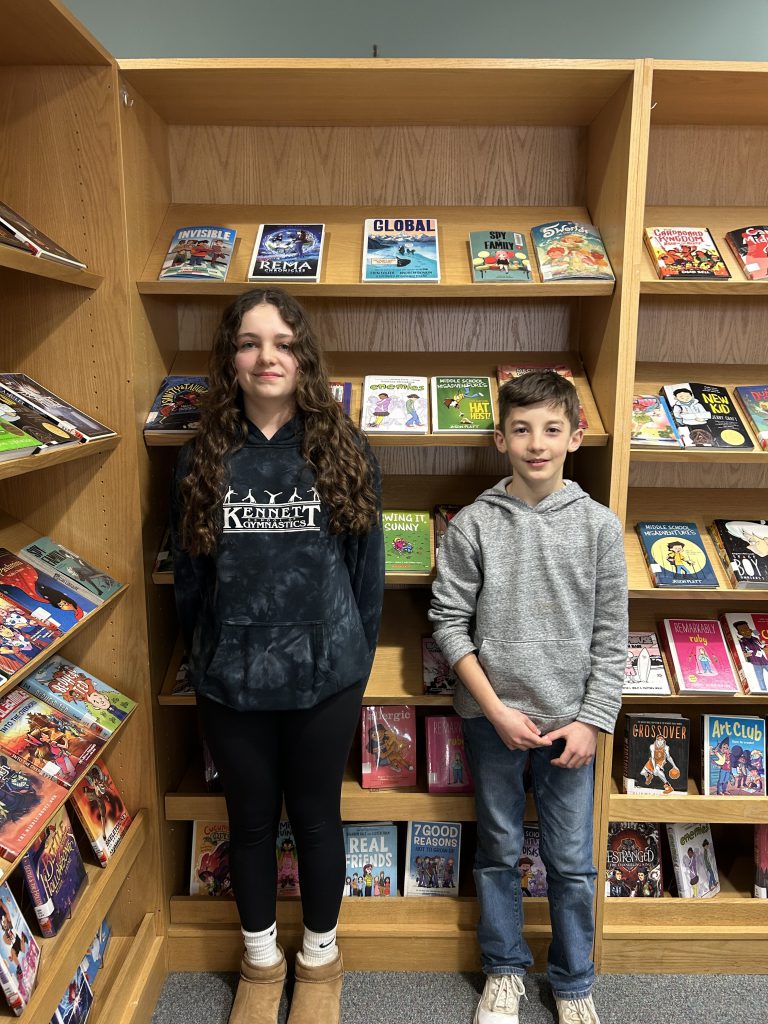 Eighth grader Cora Massaro and seventh grader Luke Hand smile for a photo in front of a books on shelves in the library. 