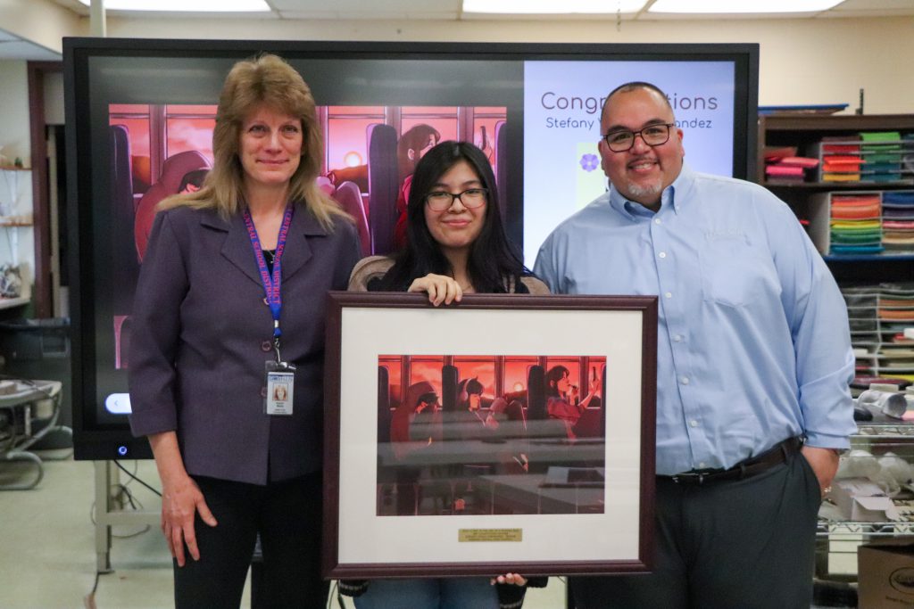 From left to right, Goshen transportation director Karen Wells (left) with artist Stefany Vidals-Hernandez (middle) and Leonard Bus Sales' Jose Fontanez (right) stand for a photo in the art room. They are posing in front of a computer screen that has a congratulations message to Stefany with her work on display. 