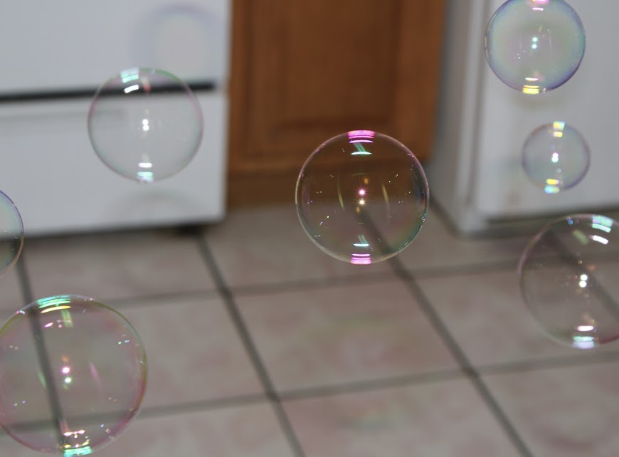 Bubbles floating in a kitchen with tole floors, a door and a fridge in the background. 