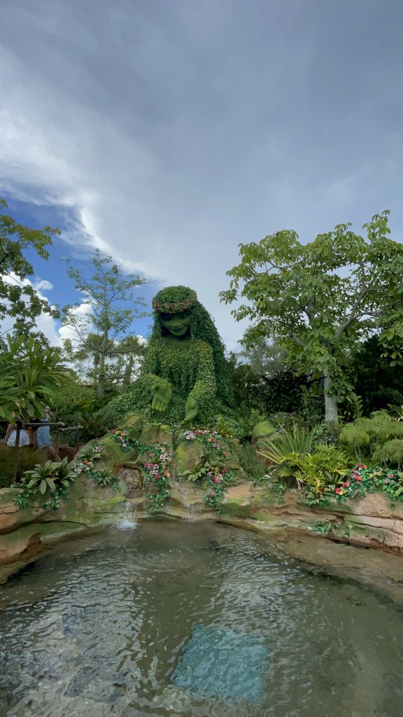 A pond with a person sculpted out of plants next to the water. There are trees and plants all around the sculpture with clouds in the sky above. 