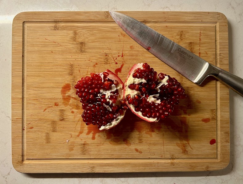 A pomegranate is cut open on a cutting board with a knife next to it covered in red juice. The seeds are still in the pomegranate and there is juice on the cutting board. 