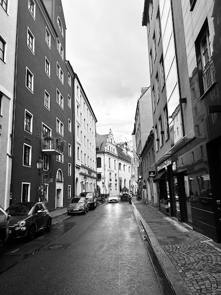 Buildings on either side of a narrow street in black and white. There are cars parked on the road and a car coming down the road. There is a brick sidewalk next to the road. 